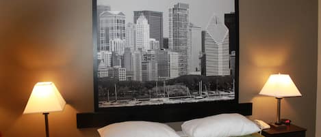 A hotel room with a large cityscape picture on the wall, two bedside lamps, a bed with two pillows, and a nightstand with a phone and a clock.