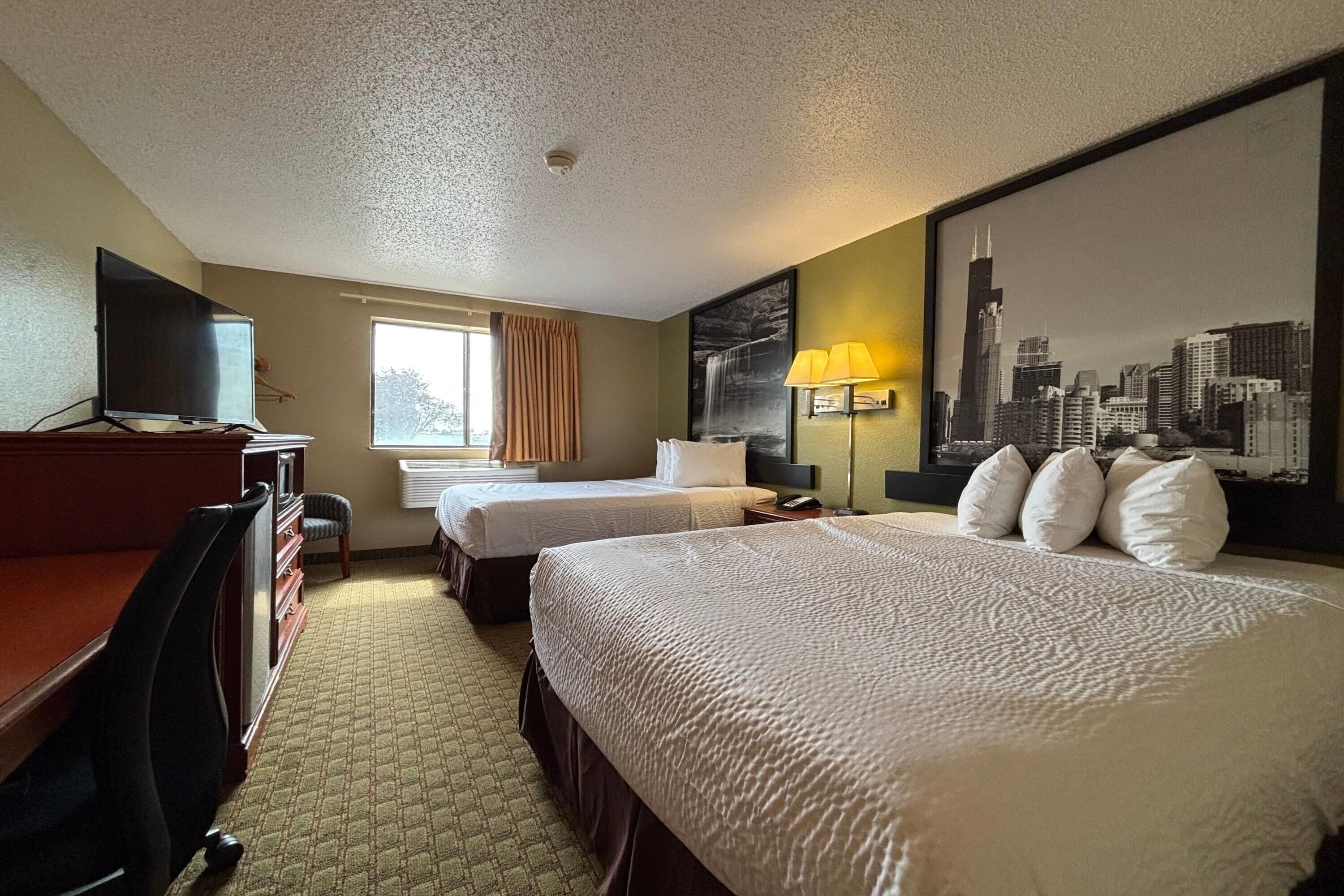 A hotel room with two beds, a desk, a television, and a cityscape painting.