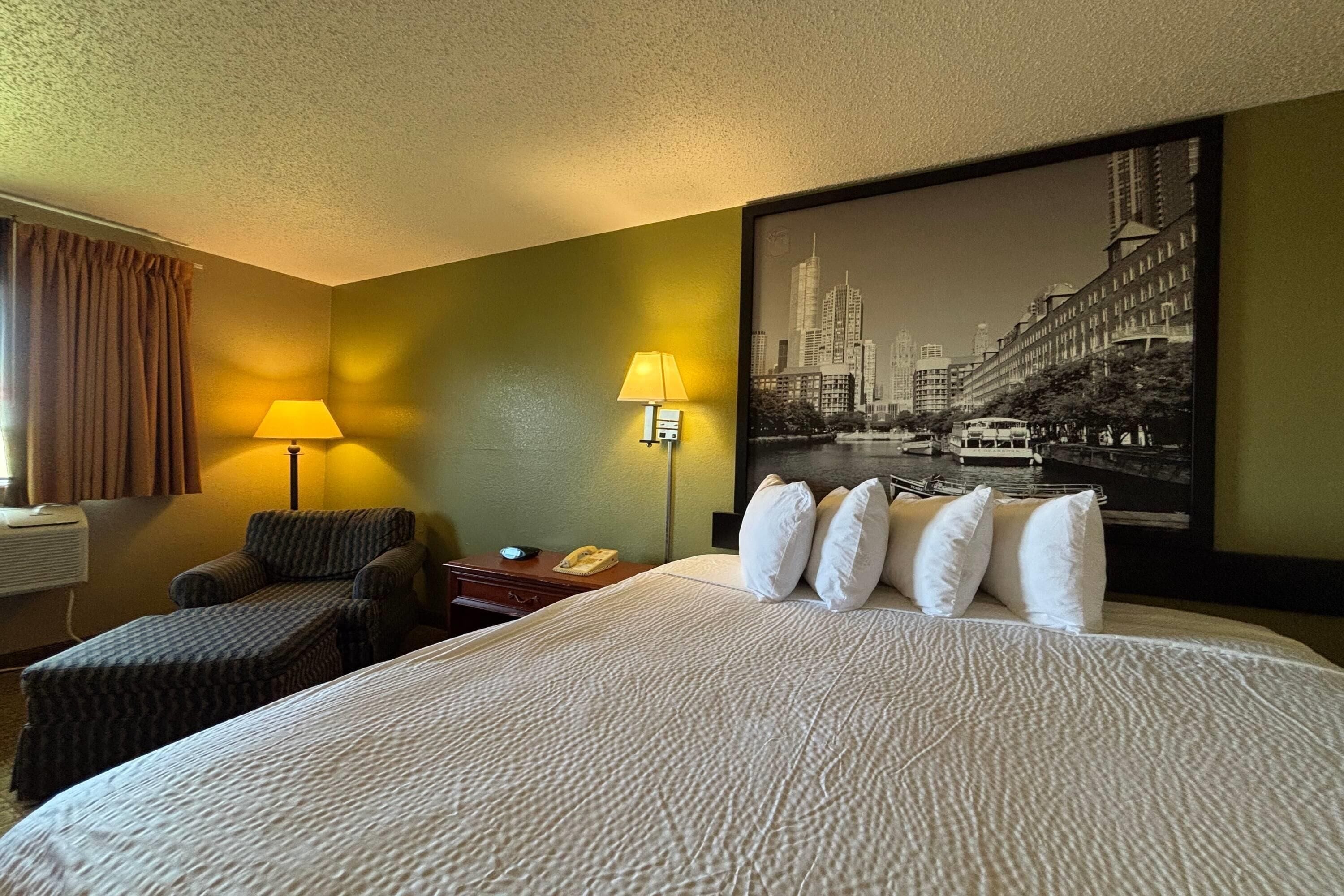 A hotel room with a large bed, two bedside lamps, a chair, a small table, and a cityscape painting on the wall.