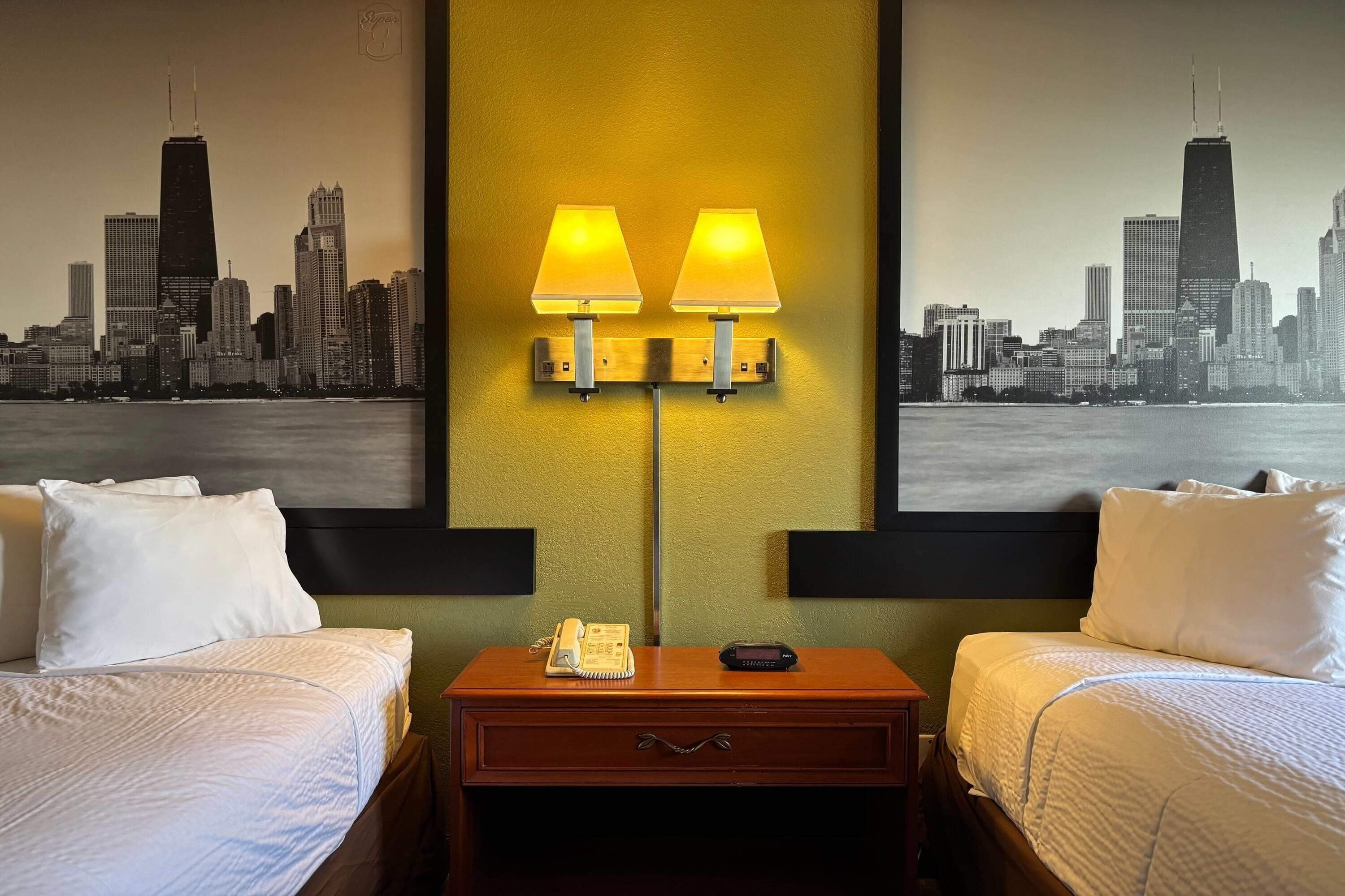 A hotel room with two beds, a nightstand with a lamp, a phone, and a digital clock. The wall features a cityscape painting.