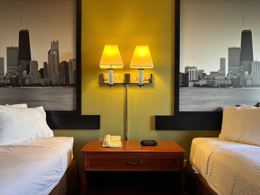 A hotel room with two beds, a nightstand with a lamp, a phone, and a digital clock. The wall features a cityscape painting.