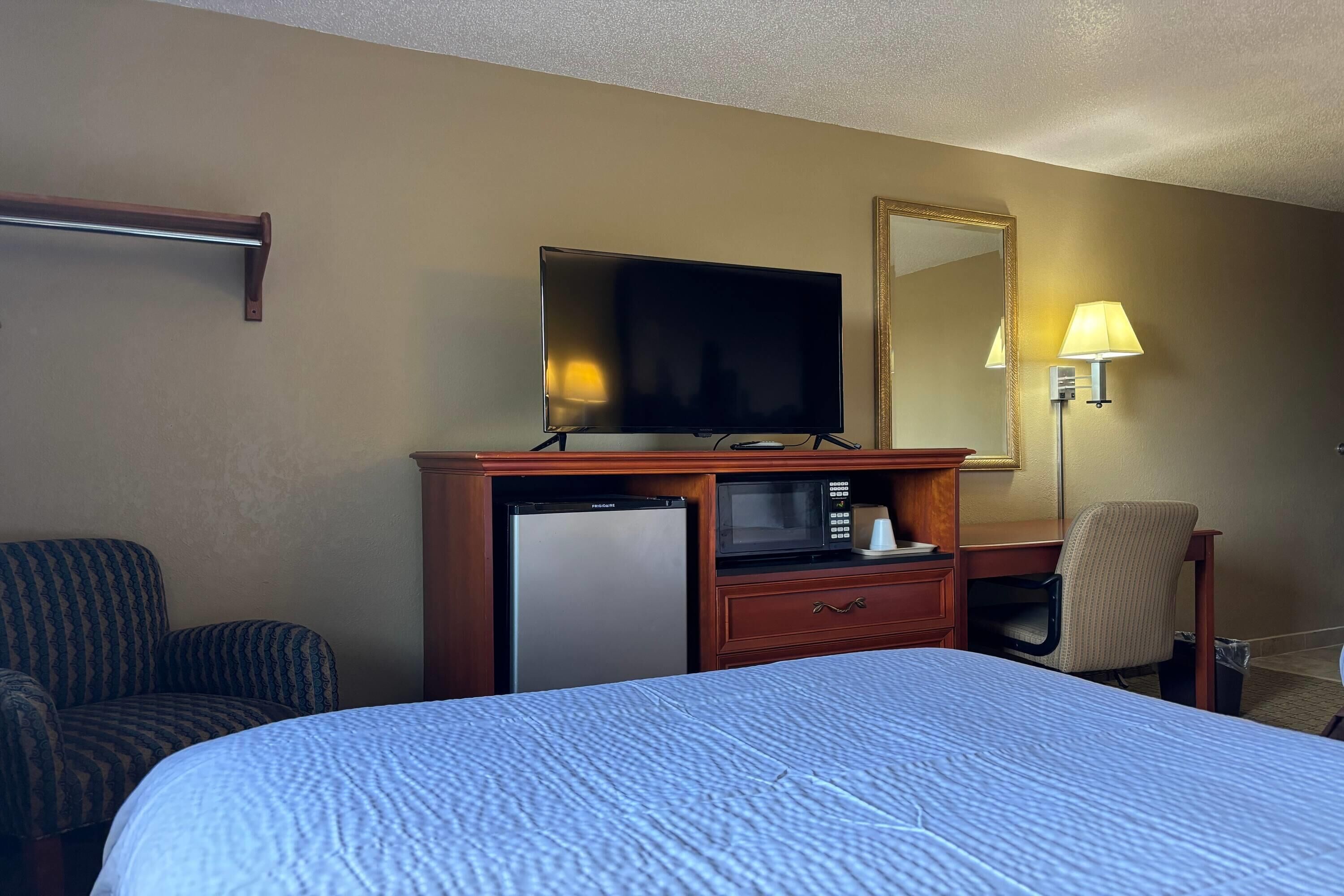 Ein Hotelzimmer mit Bett, Schreibtisch mit Fernseher, Mikrowelle, Kühlschrank, Sessel und Lampe.