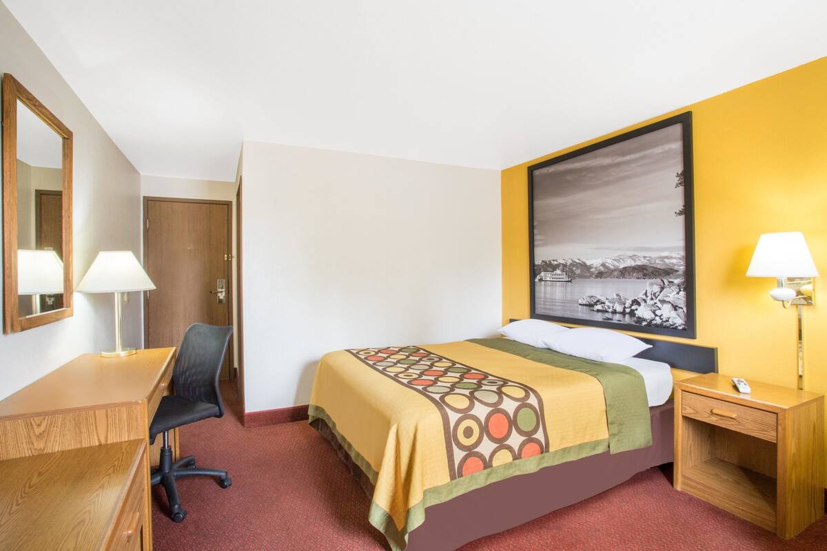 A hotel room with a bed, a desk, a chair, a nightstand, a lamp, and a large framed picture on the wall.