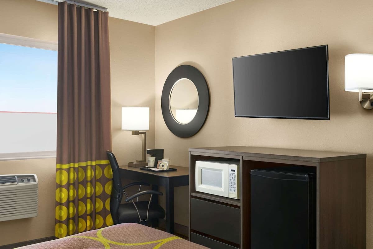 A hotel room with a bed, a desk with a lamp, a microwave, a refrigerator, a mirror, and a TV.