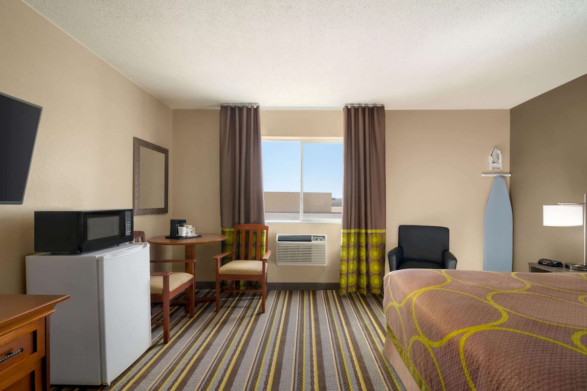 A hotel room with a bed, a desk with a microwave, a chair, a small table, an air conditioning unit, and a window with curtains.