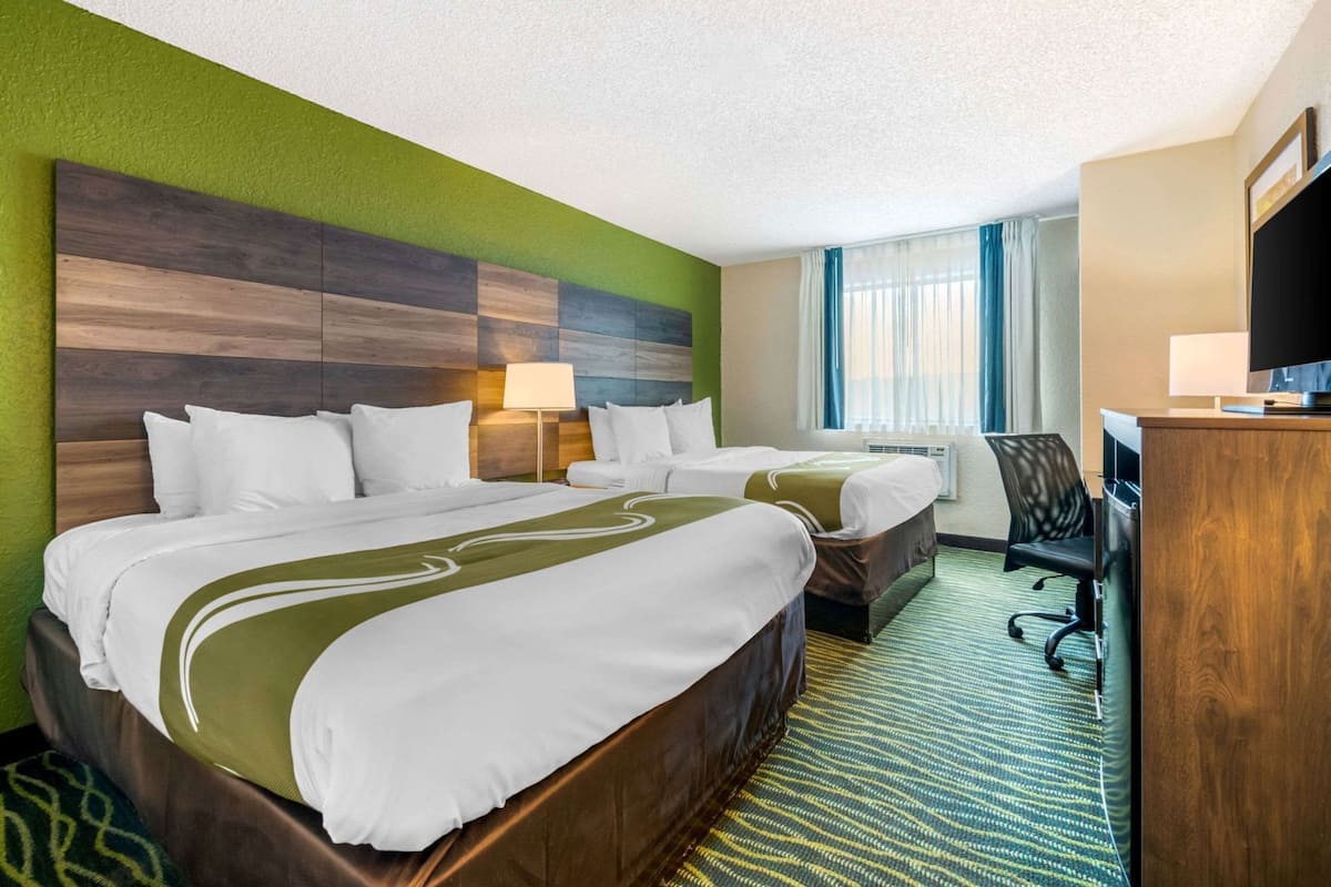 A hotel room with two beds, a desk, a TV, and a window with curtains.