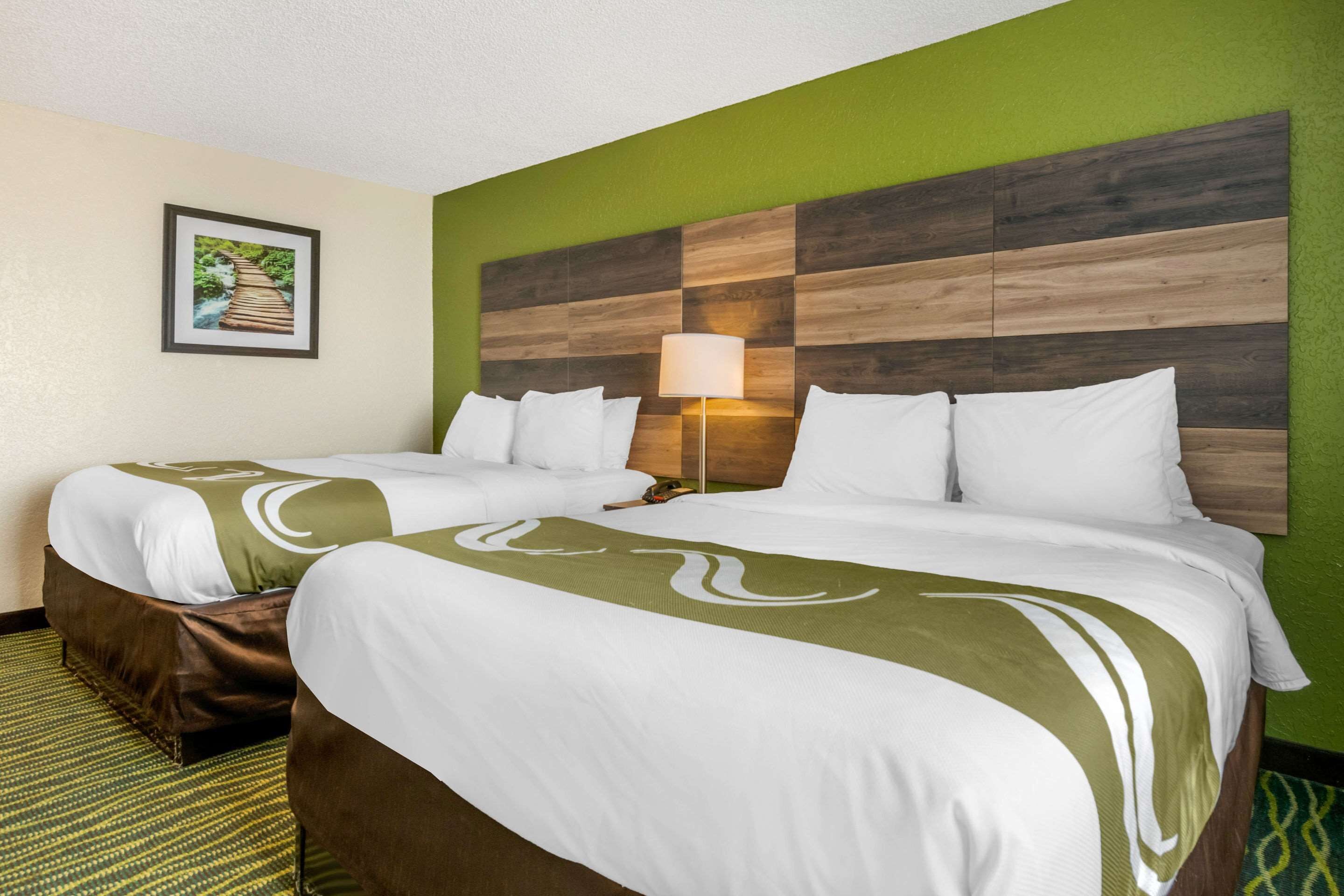 Ett hotellrum med två sängar, en sänggavel med en träpaneldesign, en inramad tavla på väggen och en lampa bredvid en av sängarna.