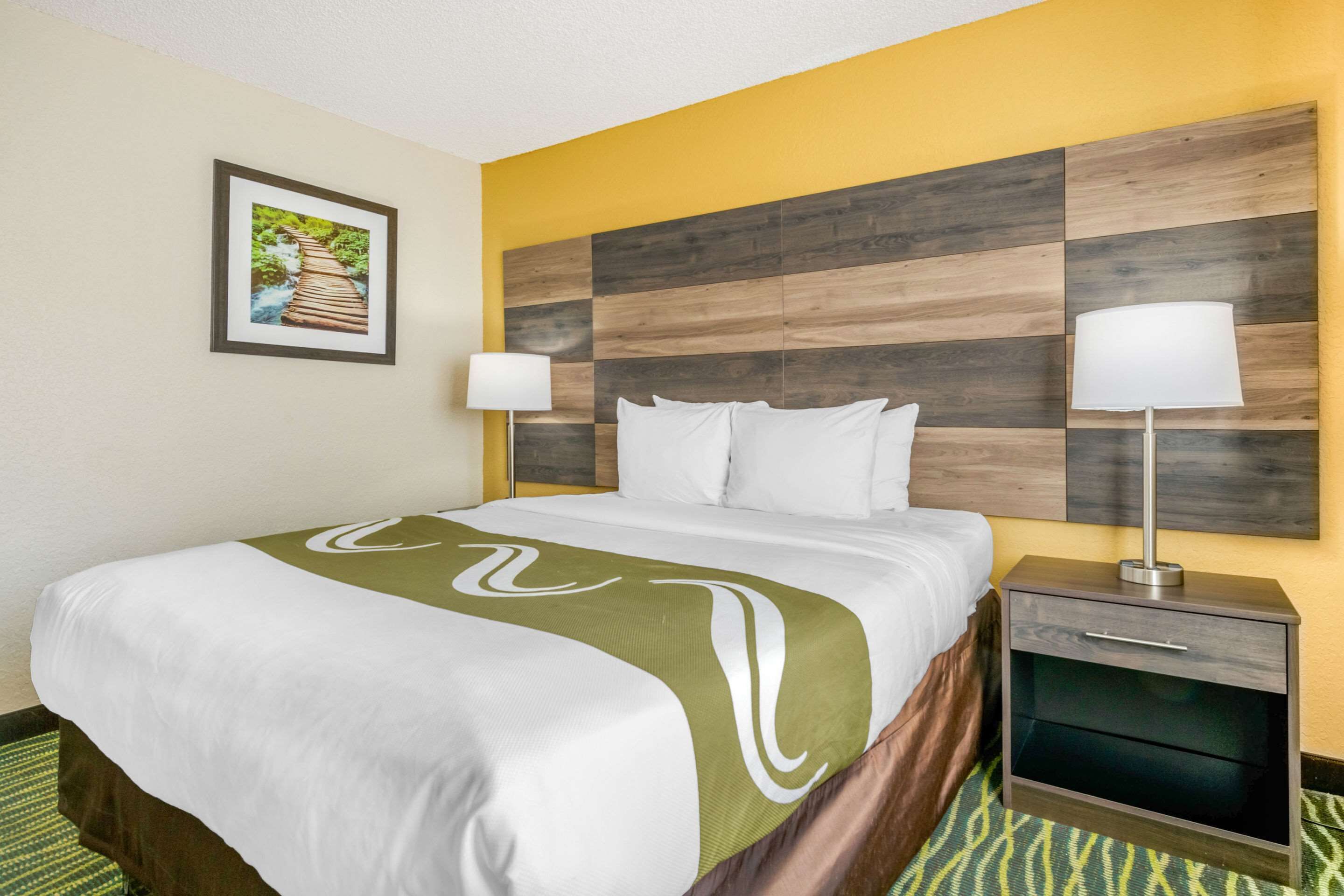A hotel room with a bed, two bedside tables with lamps, a framed picture on the wall, and a patterned carpet.