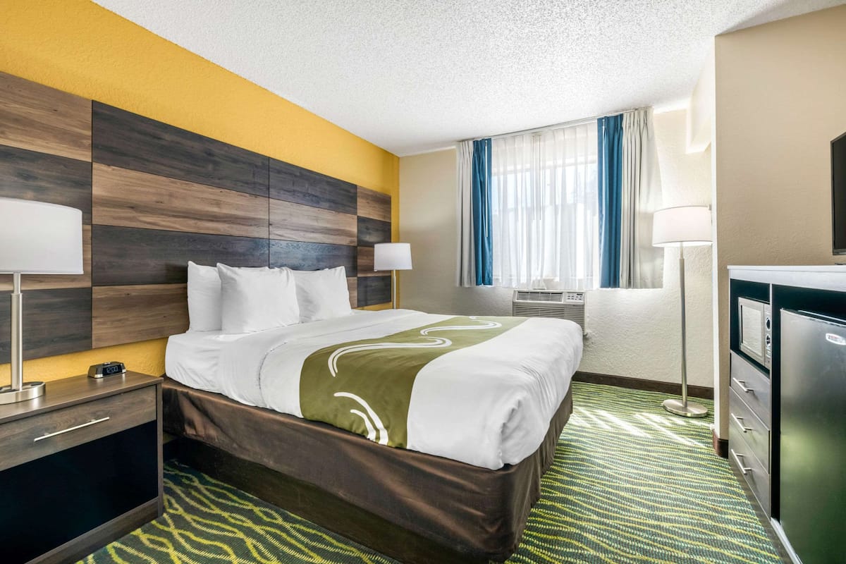 A hotel room with a bed, bedside tables, a desk, a lamp, a dresser, and a window with curtains.