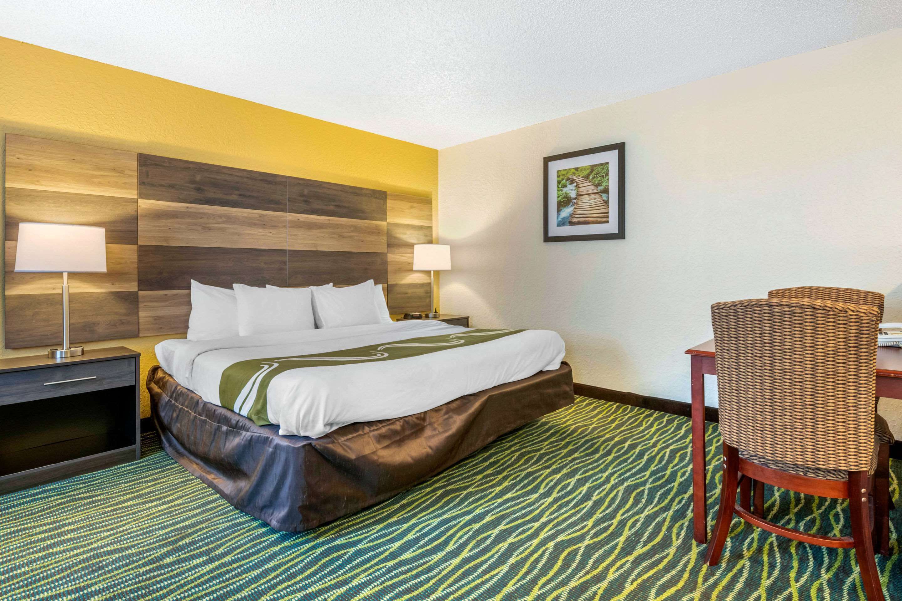 Habitación de hotel con cama, dos mesitas de noche con lámparas, una silla de mimbre y una alfombra estampada.