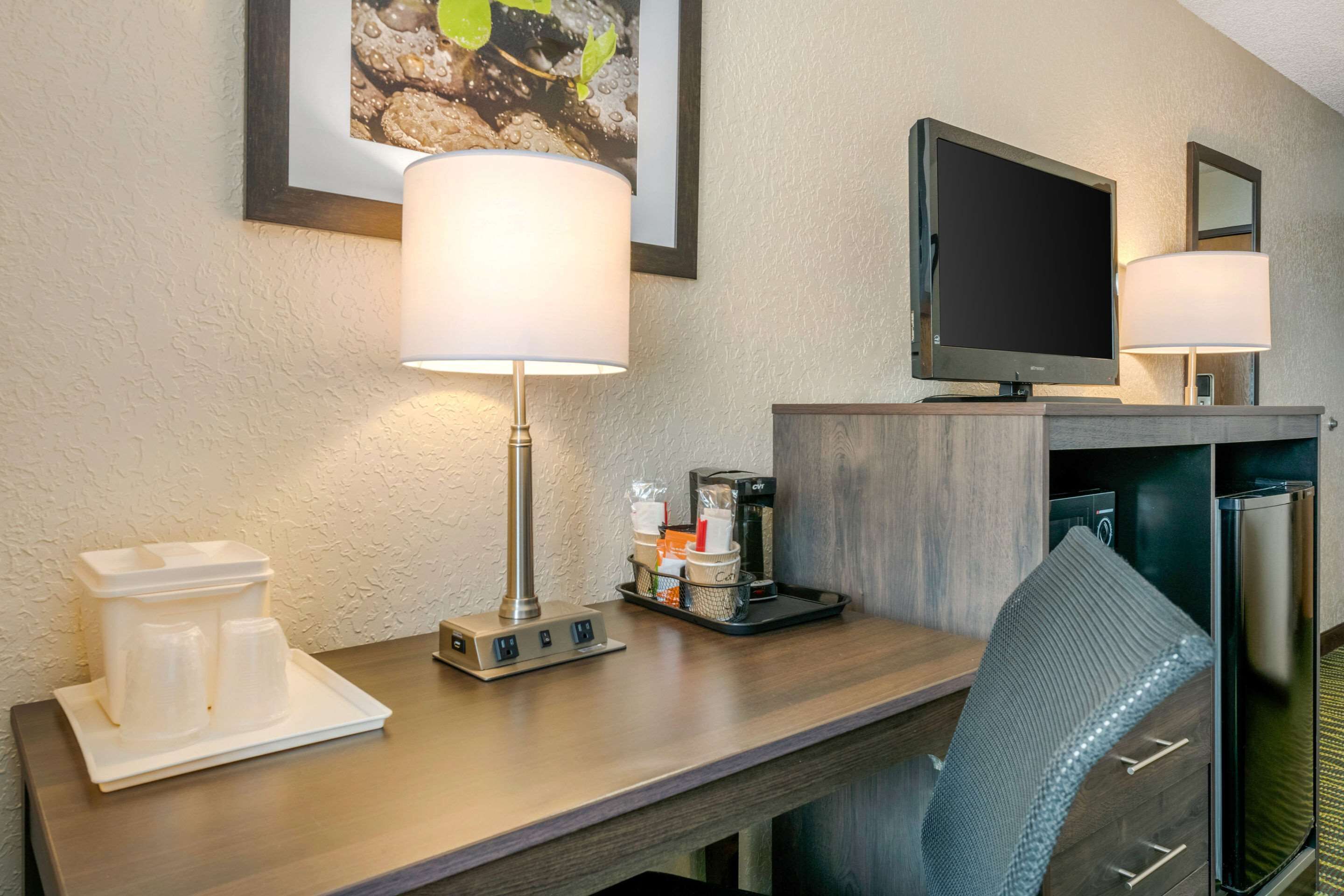 A hotel room with a wooden desk, two lamps, a television, a coffee maker, and a refrigerator.