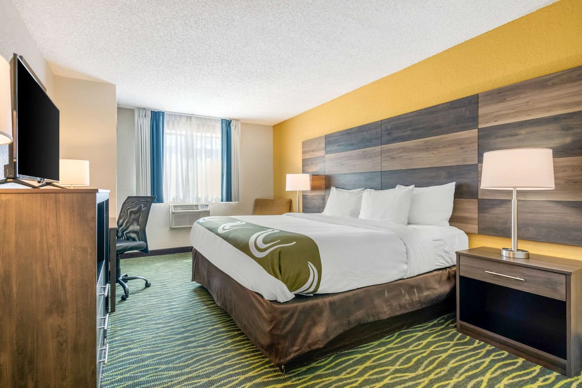 A hotel room with a bed, bedside table, lamp, desk, chair, and a TV.