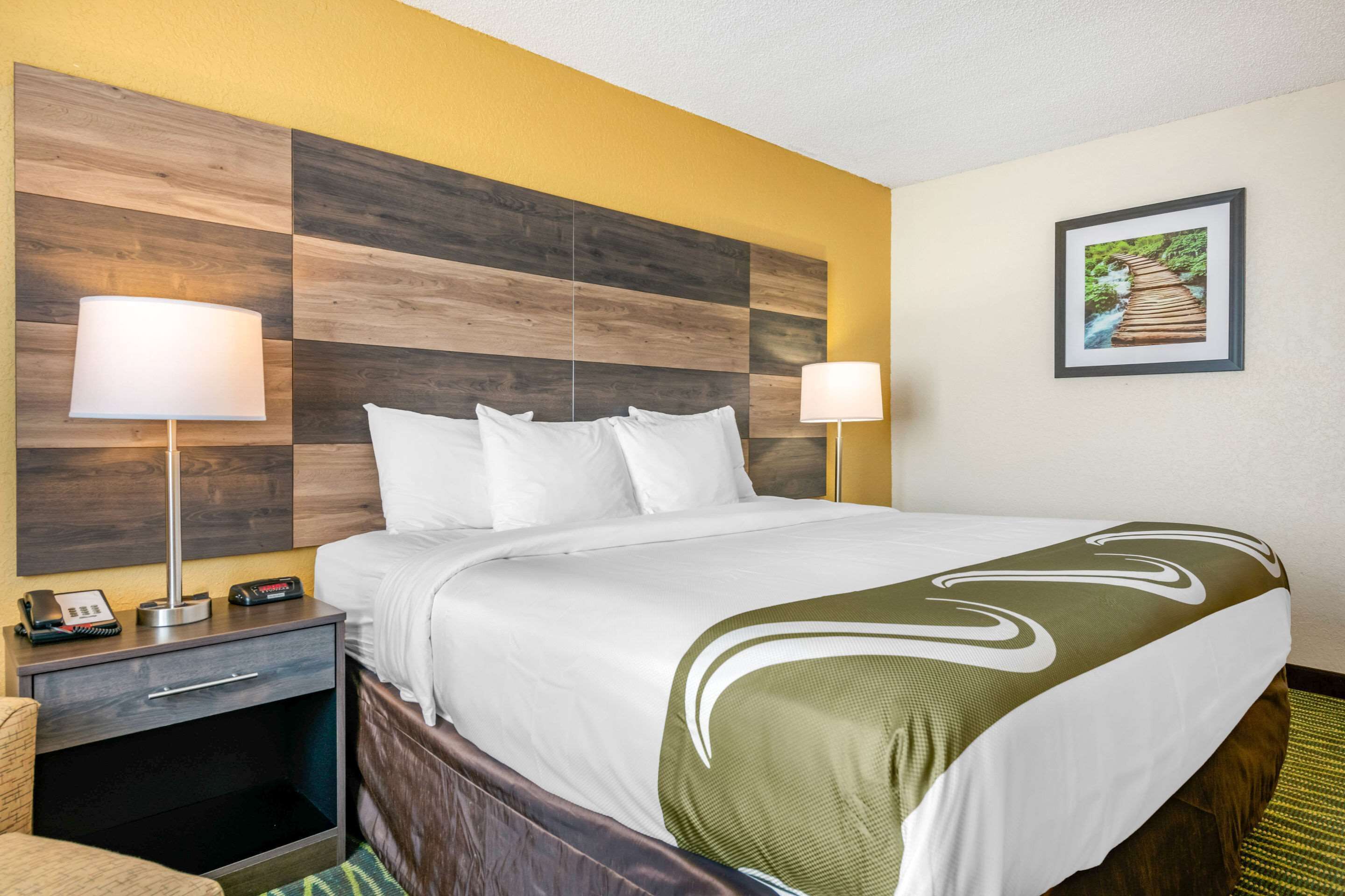 Habitación de hotel con cabecera de madera, cama con ropa blanca, dos lámparas de noche, mesita de noche con teléfono y reloj digital, y un cuadro enmarcado en la pared.
