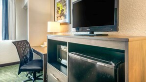 A hotel room with a flat-screen TV mounted on the wall, a small refrigerator, a desk with a chair, and a window with curtains. - Quality Inn & Suites Leesburg Chain of Lakes (Leesburg)