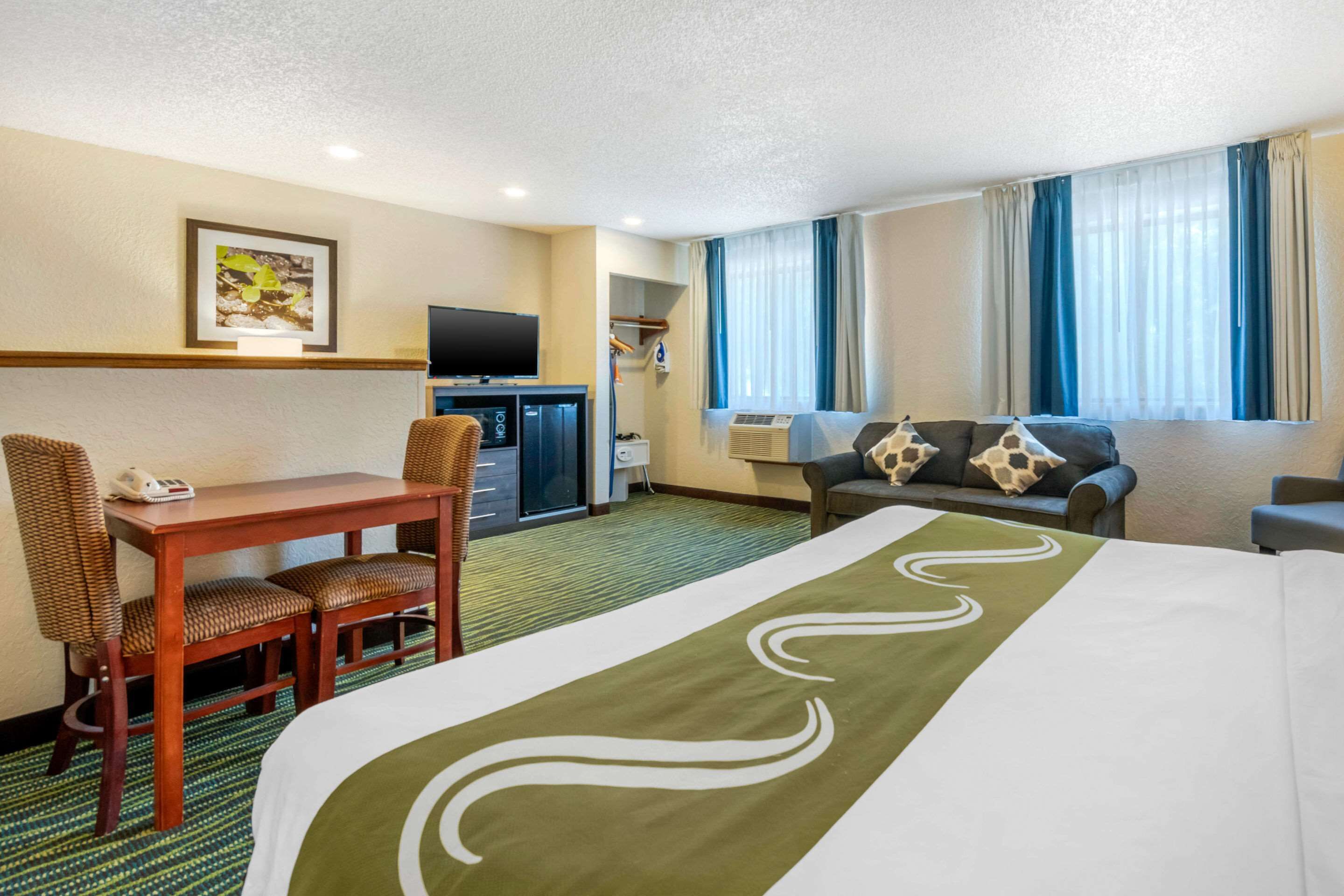 Quarto de hotel com cama, escrivaninha com cadeiras, televisão e sofá.