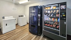 A room with a vending machine labeled 'COLD drinks' and a 'SNACKS' vending machine, a washing machine, and a dryer. - Motel 6 Ennis, TX (Ennis)