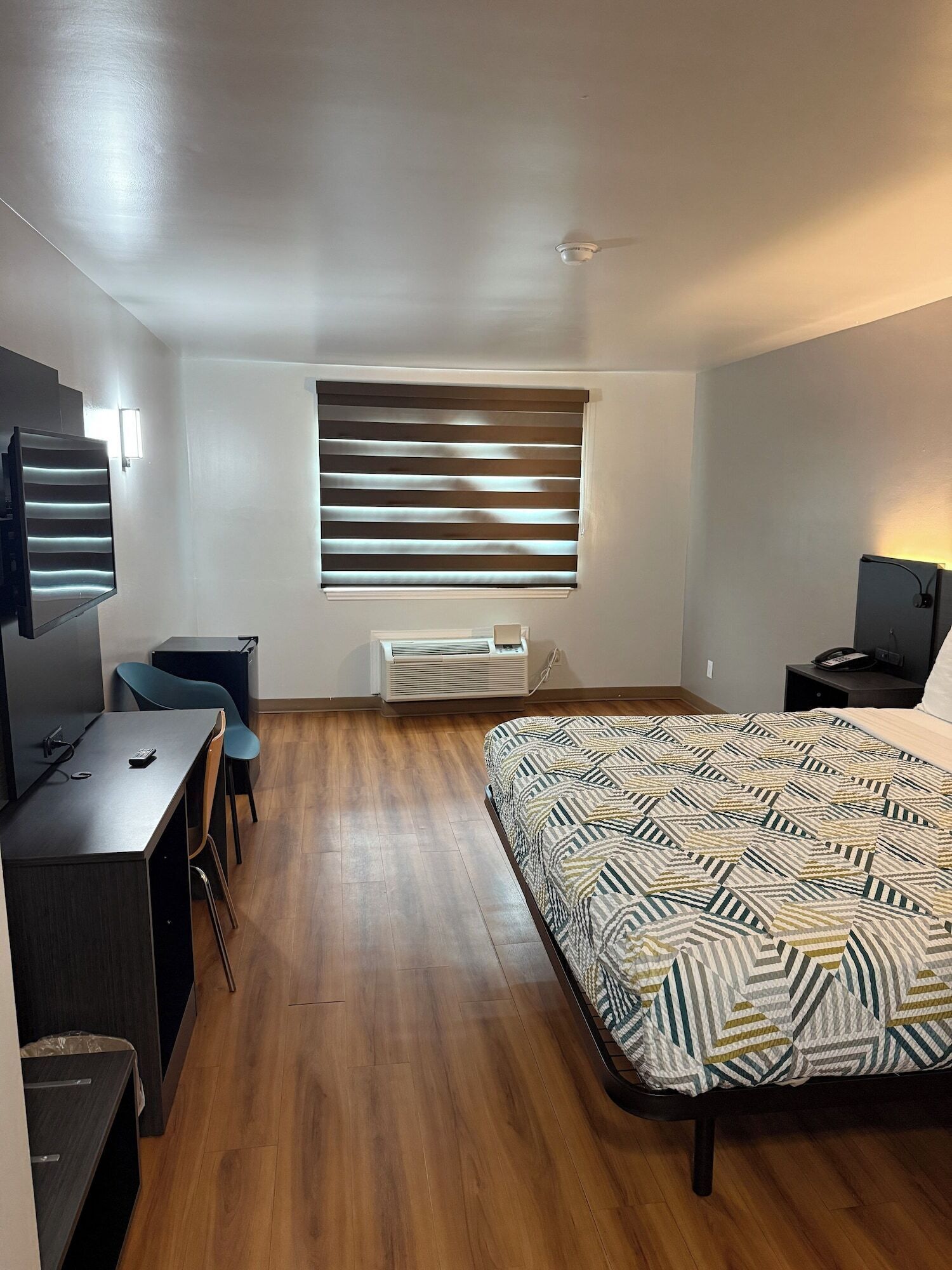 Un dormitorio con cama, escritorio, silla, ventana con persianas, ventilador de techo y unidad de aire acondicionado.