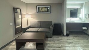 A hotel room with a sofa, a coffee table, a lamp, a framed picture, and a bathroom with a sink and mirror. - Quality Inn & Suites at Airport Blvd I-65 (Mobile)