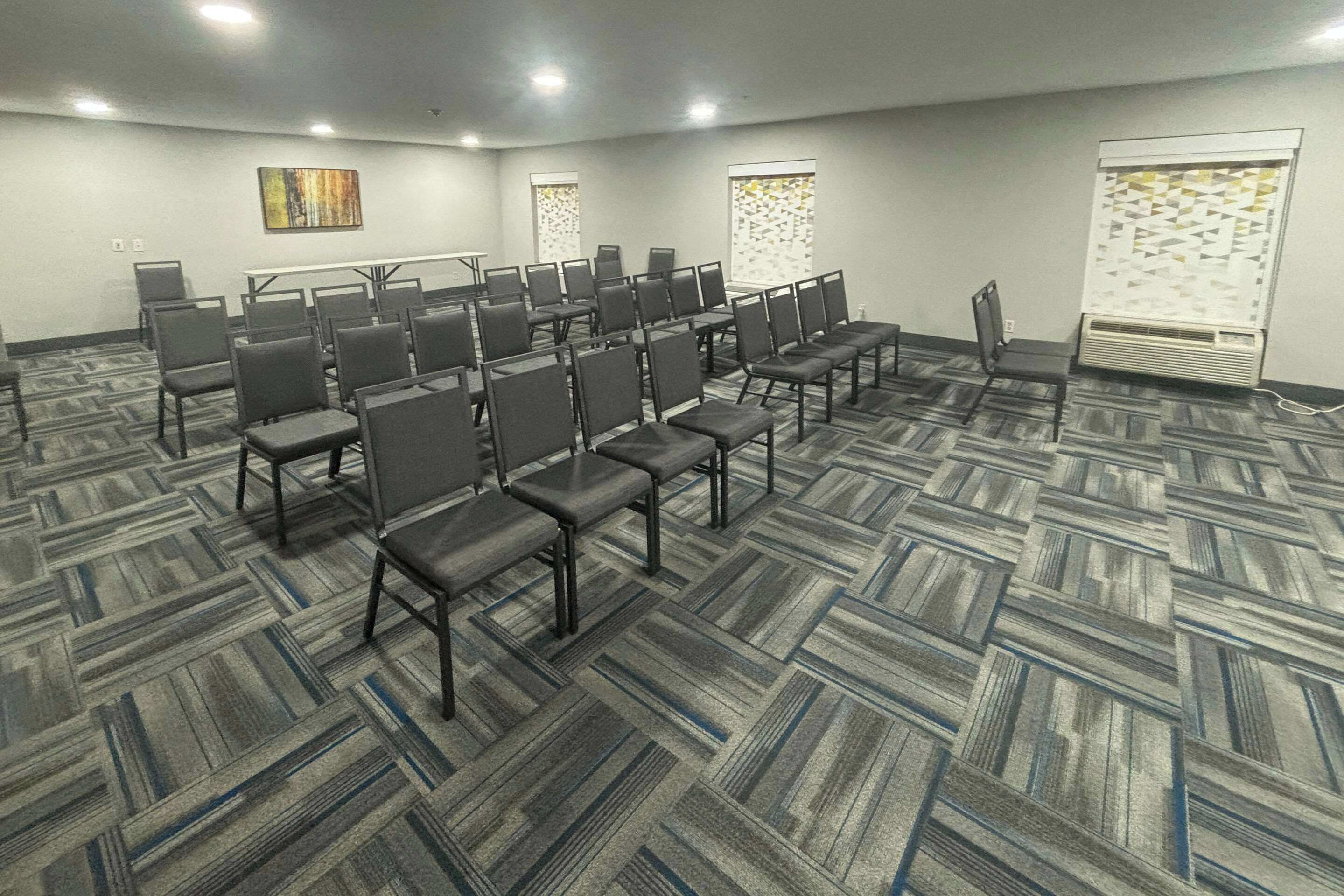 Un salón de conferencias con filas de sillas, piso alfombrado y un amplio ventanal con persianas.