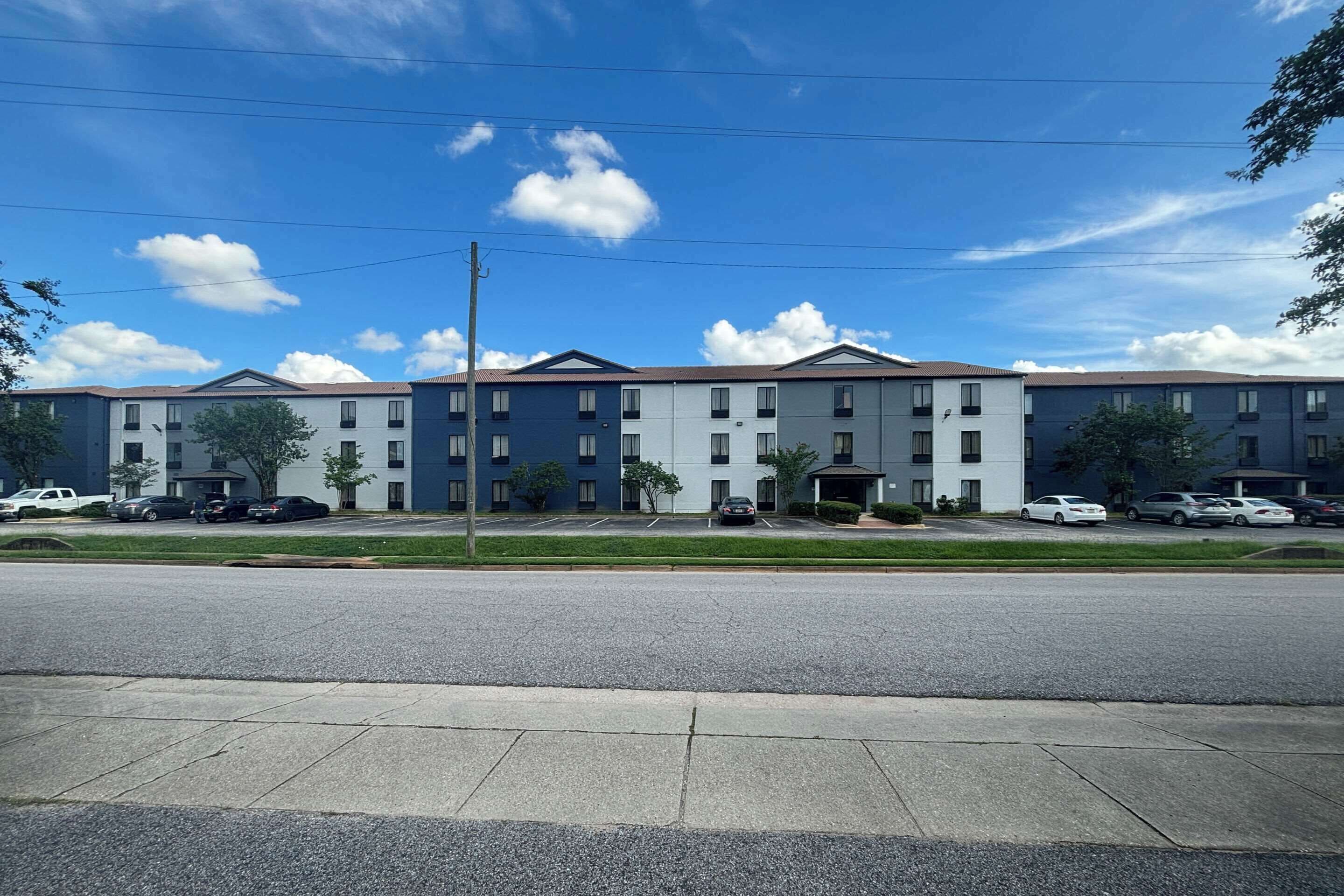 Ein mehrstöckiges Hotel mit einer Reihe von Fenstern auf jeder Etage, einem Parkplatz davor und blauem Himmel mit Wolken.