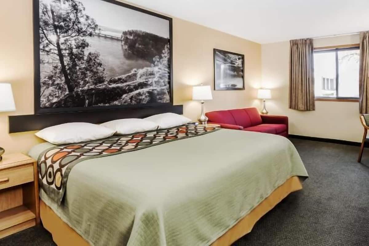 A hotel room with a bed, a desk, a chair, a red sofa, two lamps, a large framed picture, and a window with curtains.