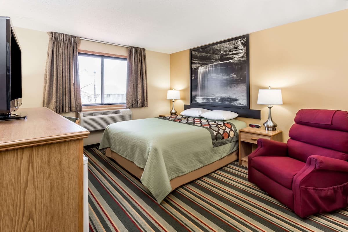 A hotel room with a bed, a desk, a chair, a TV, a window with curtains, a wall-mounted lamp, and a large framed picture.