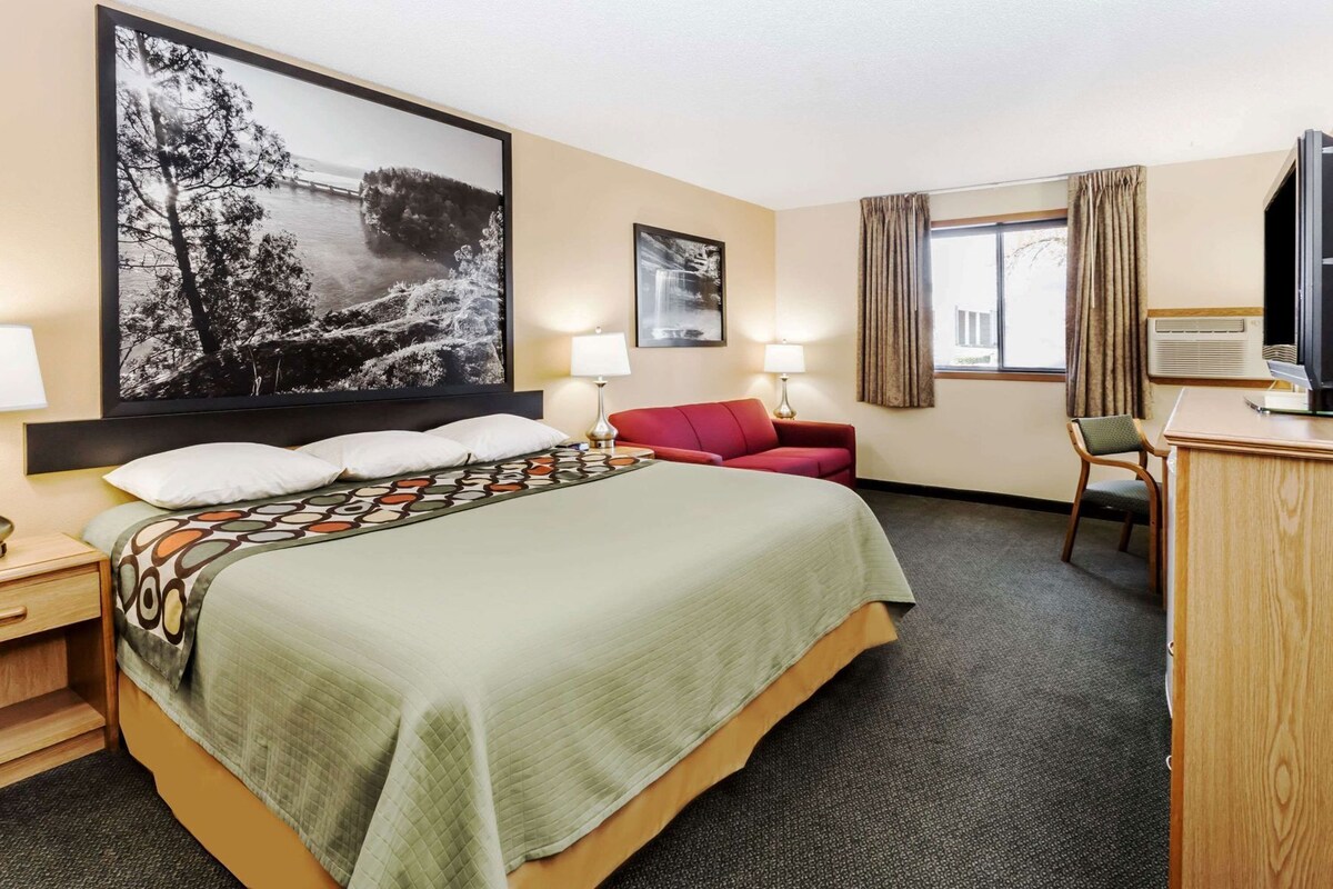 Habitación de hotel con una cama grande, un escritorio, una silla, un sofá rojo, dos lámparas y un cuadro grande en la pared.