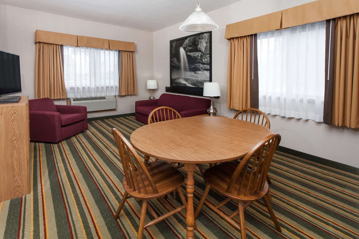 A hotel room with a wooden dining table, two wooden chairs, a red sofa, a TV, and a large window with curtains.