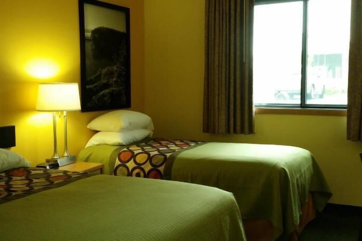 A hotel room with two beds, a nightstand with a lamp, a painting on the wall, and a window with curtains.