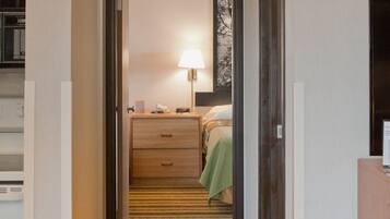 Un pasillo de hotel con piso alfombrado, una cama con cubrecama verde, una mesita de noche con lámpara y un aparador de madera.