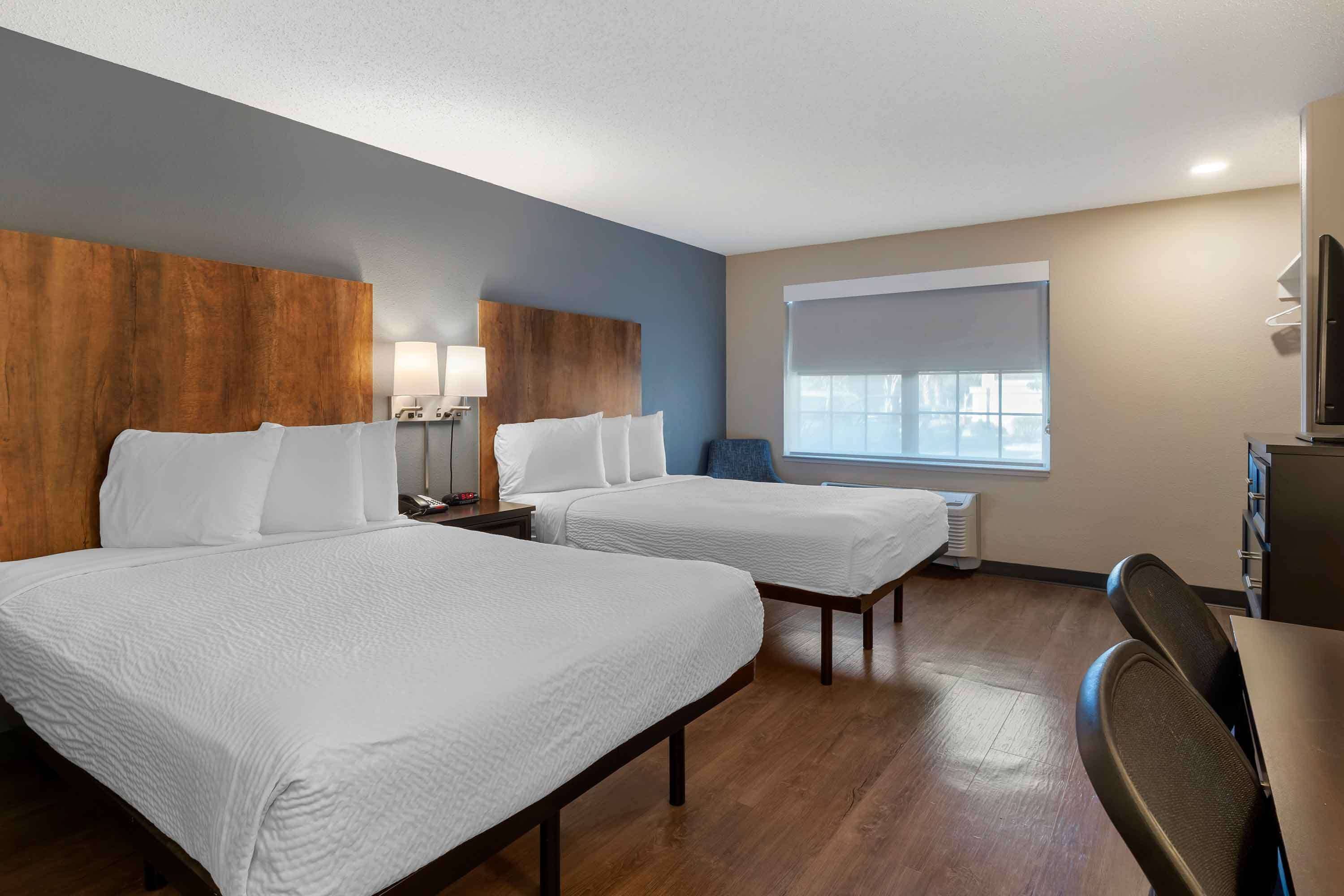 A hotel room with two beds, a desk, a chair, a window with blinds, and a wall-mounted TV.