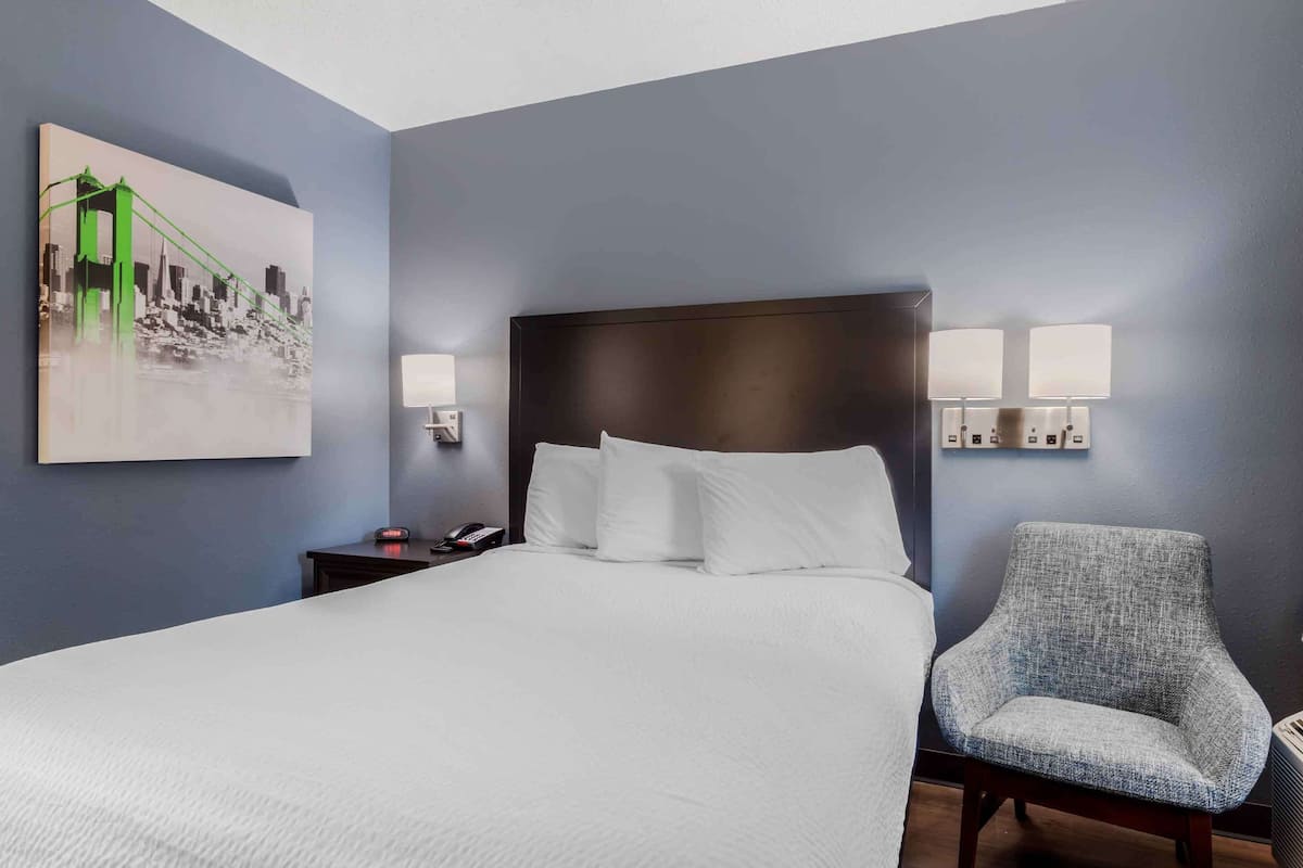A hotel room with a large bed, a nightstand with a lamp, a chair, and a painting on the wall.