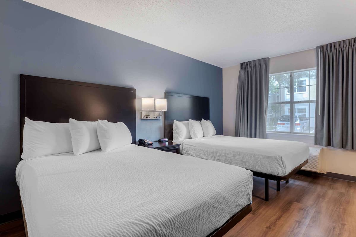 A hotel room with two beds, a dark headboard, bedside lamps, a window with curtains, and a view of the outside.