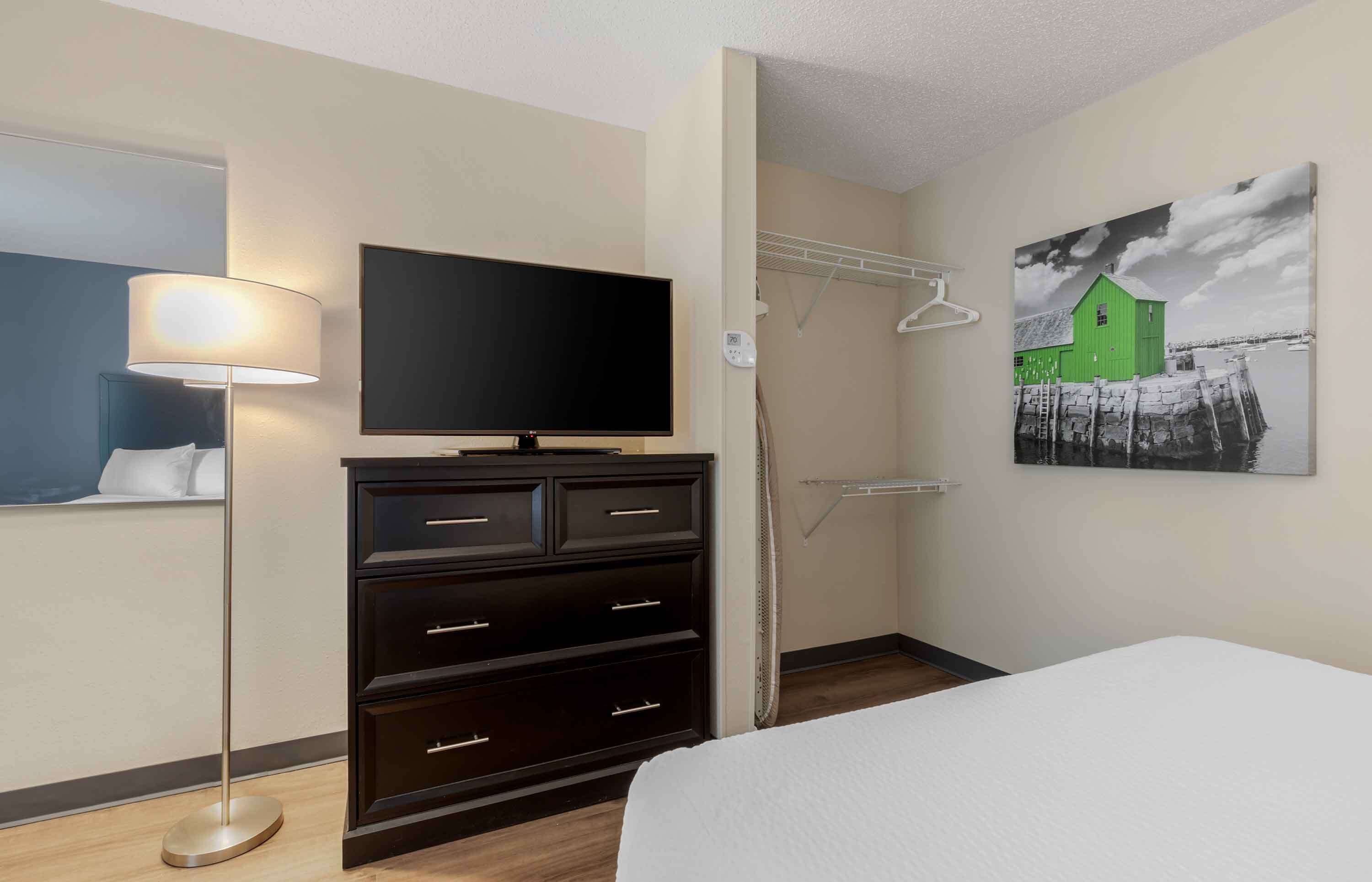 A hotel room with a bed, a flat-screen TV on a dresser, a lamp, a painting of a green house, and a closet with a hanging rack.
