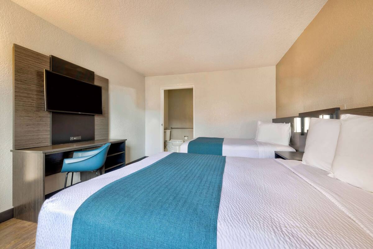 A hotel room with two beds, a desk with a chair, a flat-screen TV, and a bathroom visible through an open door.