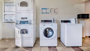 A laundry area with three stacked washers and dryers, a soap and bleach softener dispenser, and a microwave on a shelf.