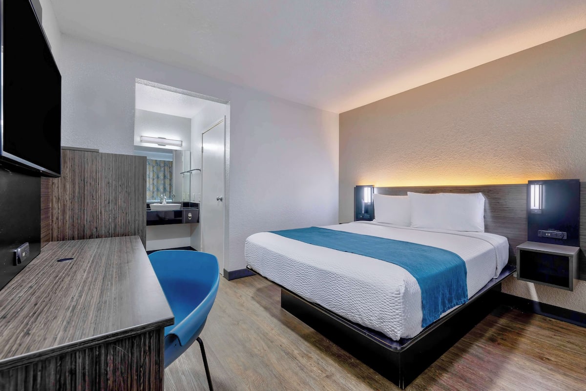 A modern hotel room with a large bed, a desk, a chair, a television, and a bathroom visible through an open door.