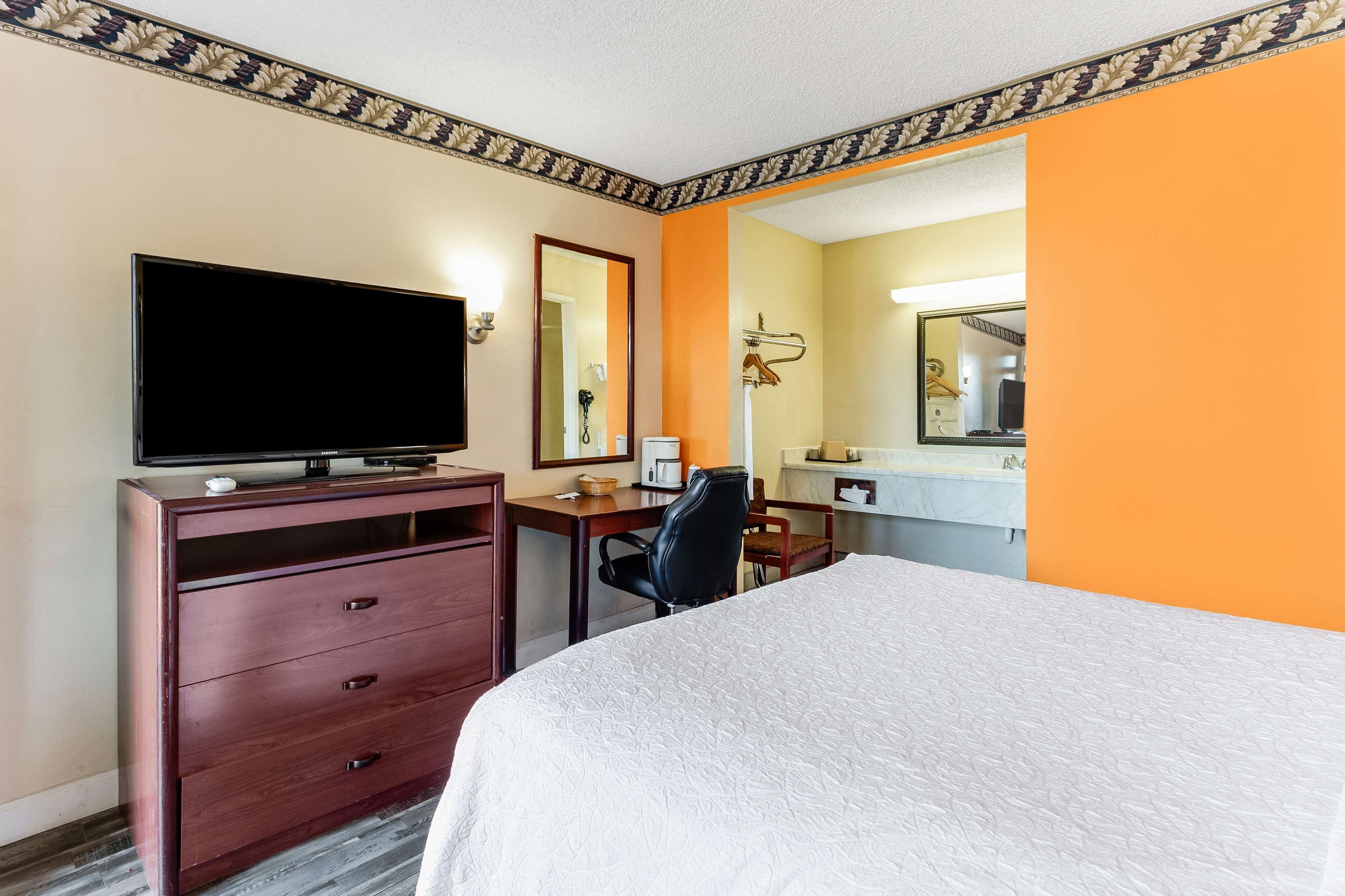 A hotel room with a bed, a desk with a chair, a television, a dresser, and a bathroom with a mirror.