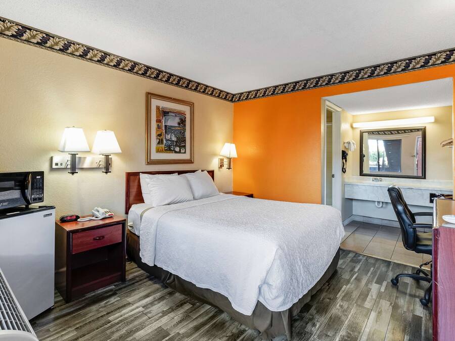 A hotel room with a bed, bedside lamps, a desk with a chair, a microwave, a refrigerator, and a mirror.
