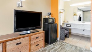 Habitación de hotel con televisor de pantalla plana, mini refrigerador, microondas, cómoda de madera y baño con espejo, lavamanos y toallero.