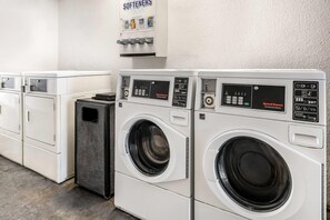 Una hilera de lavadoras industriales blancas con paneles de control, un dispensador de suavizante instalado en la pared y un cubo de basura.