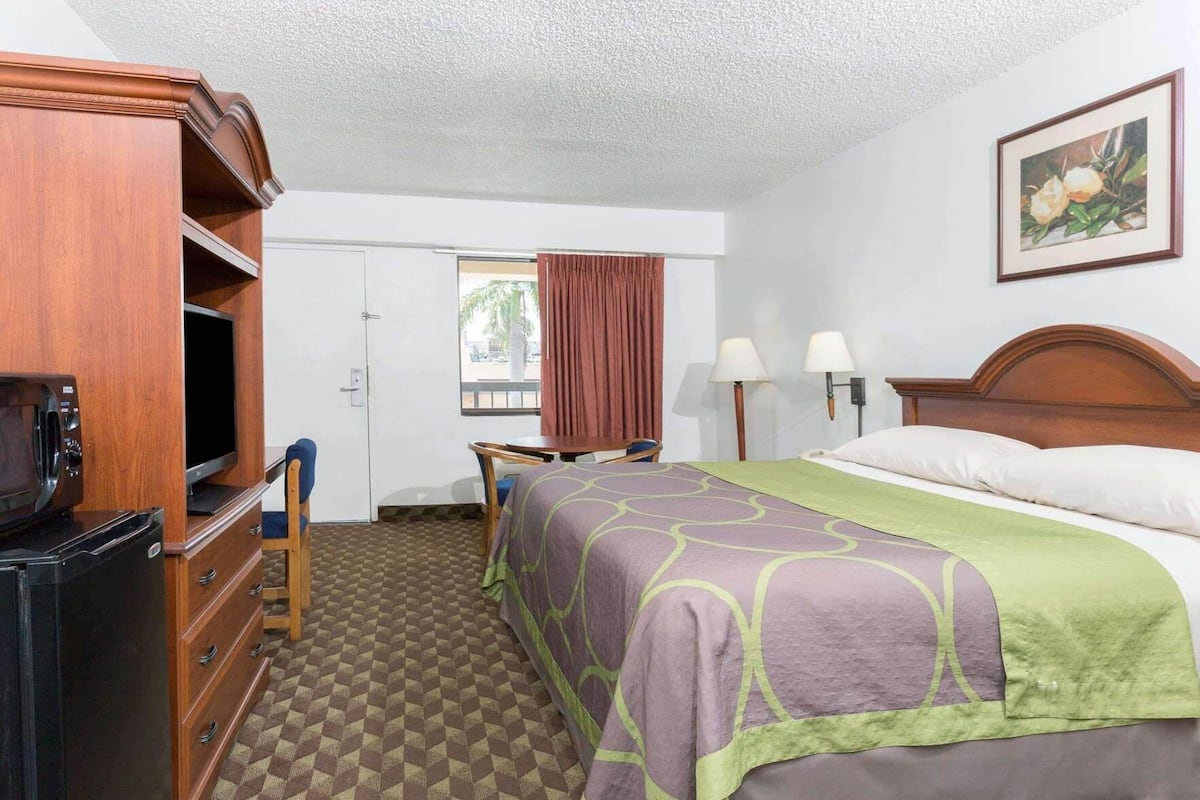 A hotel room with a bed, a desk with a chair, a television, a microwave, and a painting on the wall.
