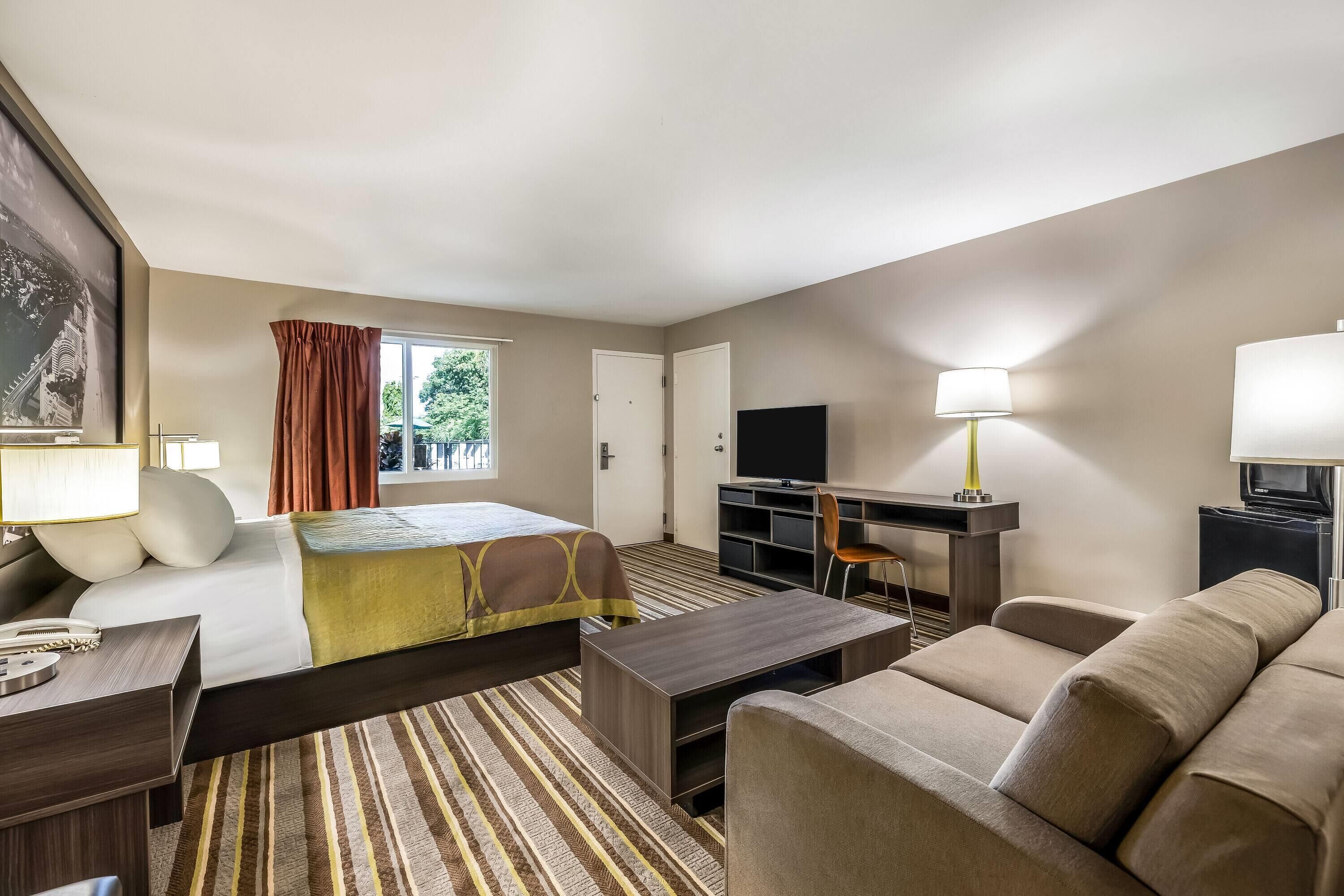 Quarto de hotel com cama, sofá, secretária e televisão, além de janela com cortinas.