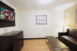 A room with a flat-screen TV mounted on the wall, a dark wooden dresser with drawers, a black leather sofa, a wooden coffee table, and a framed picture on the wall. - Red Roof Inn Morgan City (Morgan City)