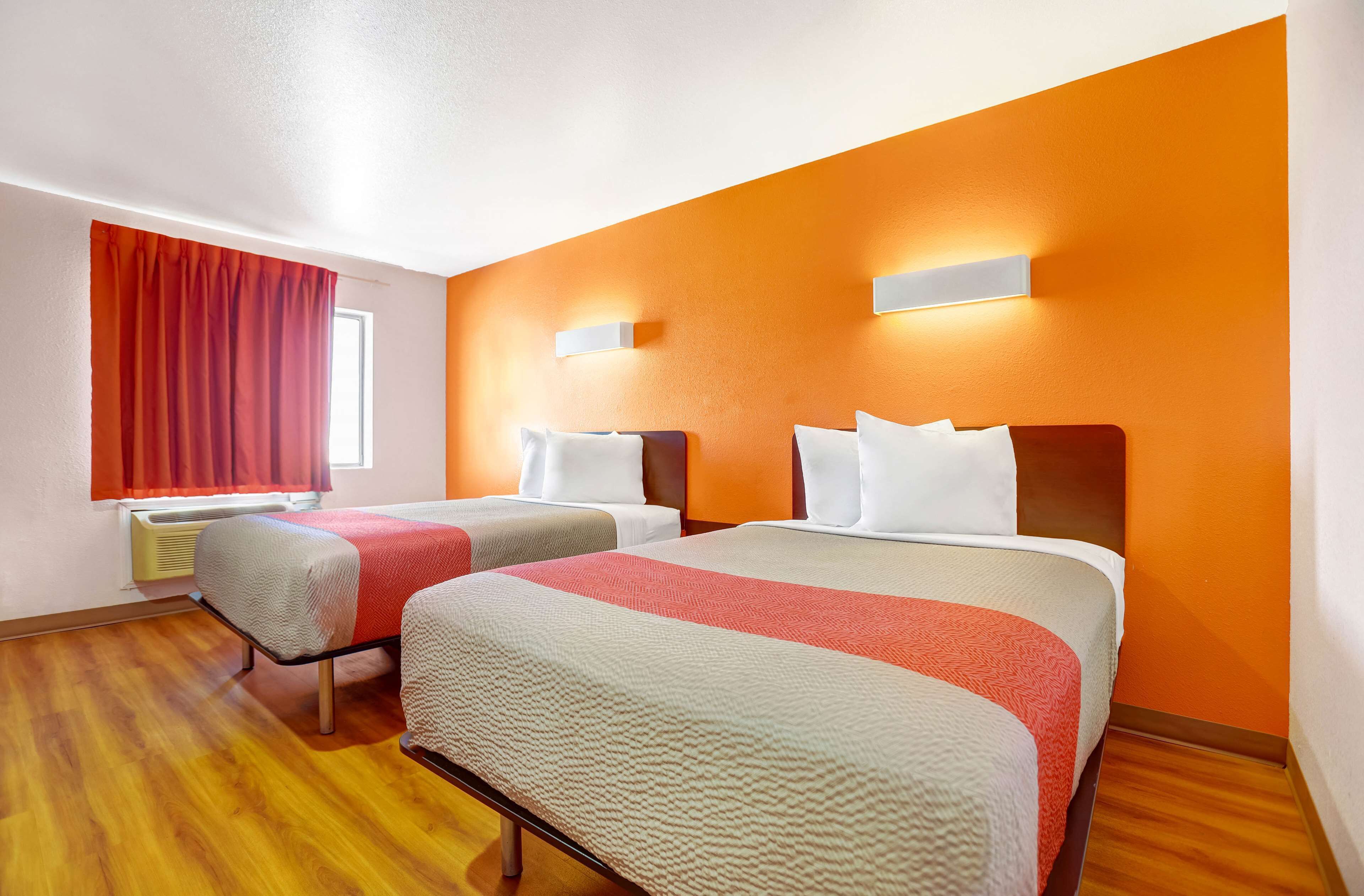 Habitación de hotel con dos camas, una ventana con cortinas rojas y una pared de acento naranja.