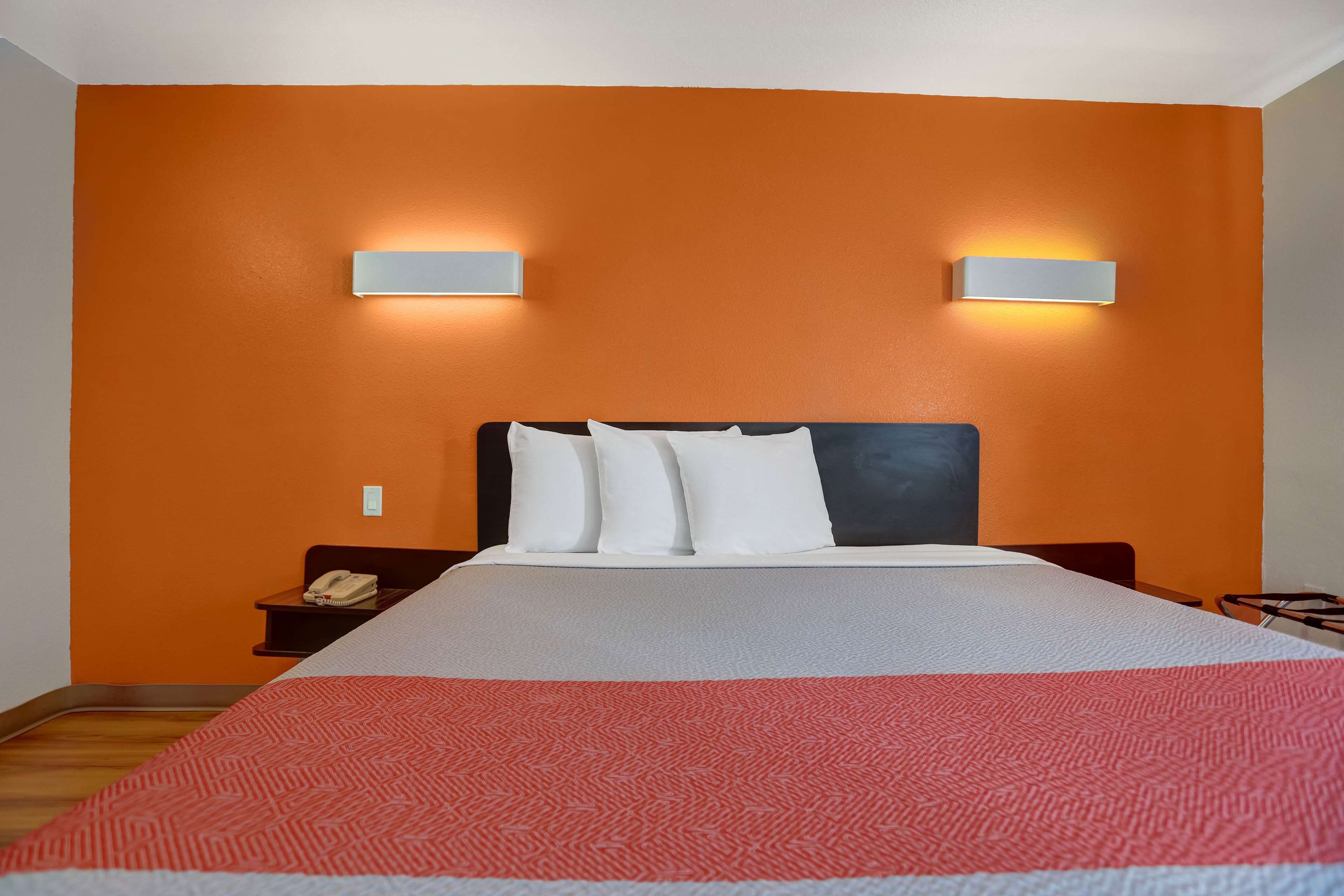 Une chambre d’hôtel avec un mur orange, un grand lit avec une literie blanche et deux appliques murales.