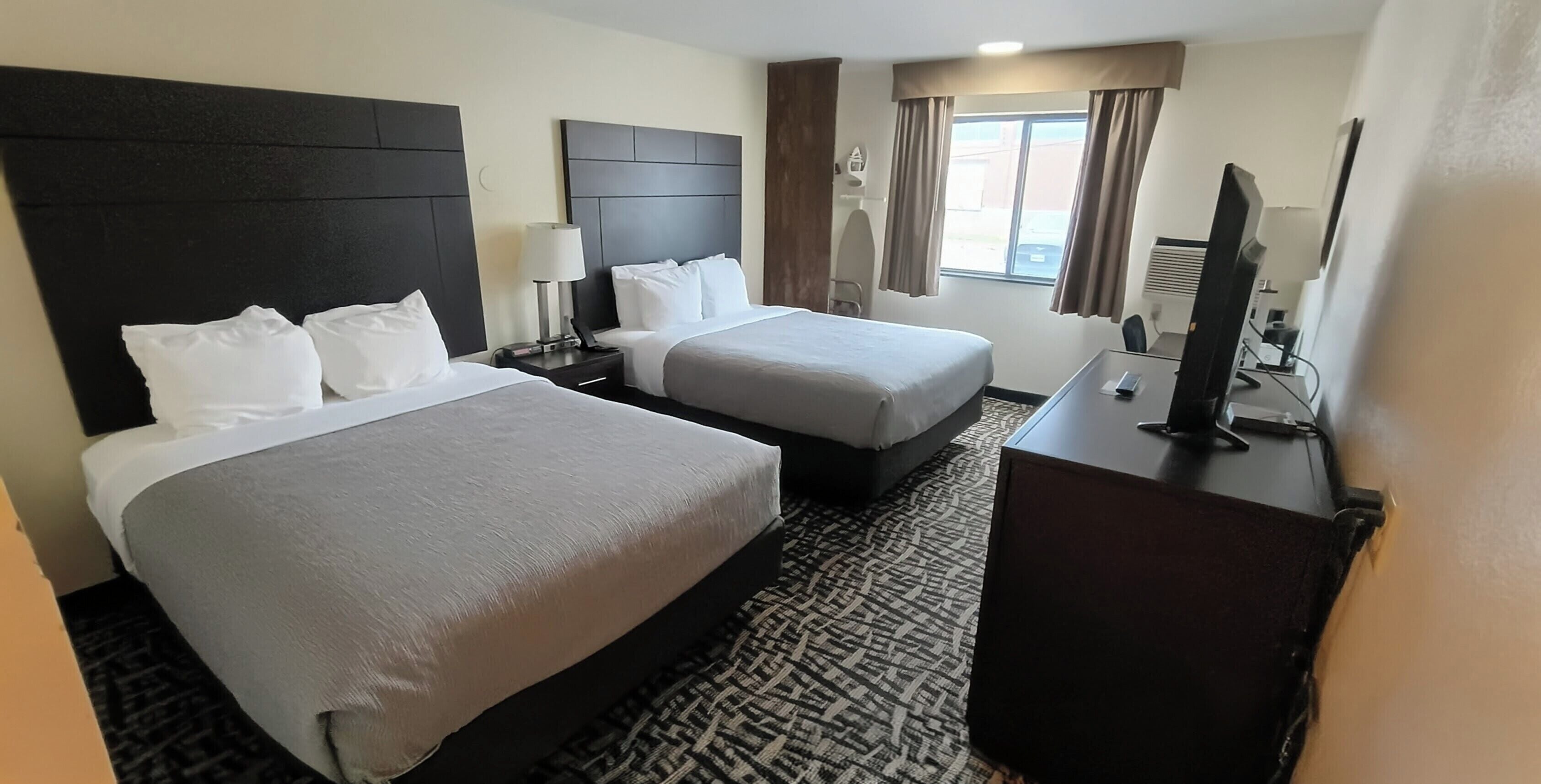 A hotel room with two beds, a desk, a TV, and a window with curtains.