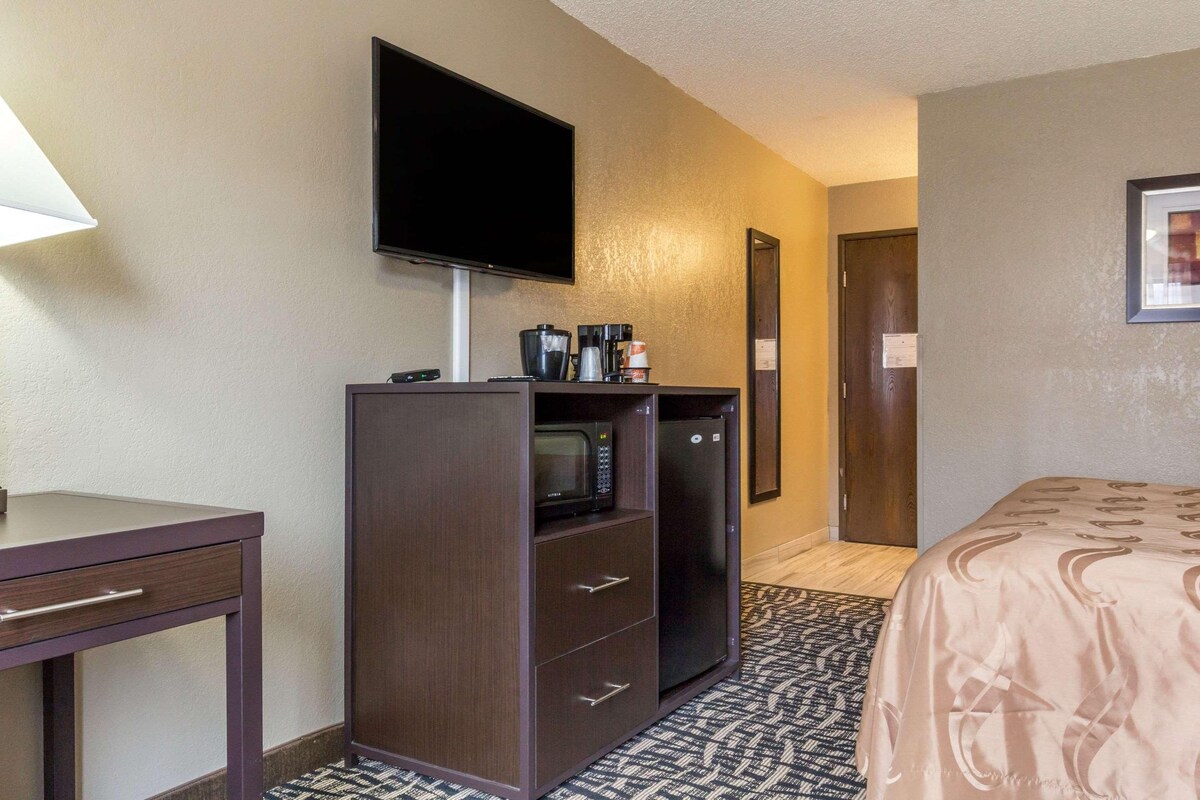 Quarto de hotel com cama, escrivaninha com luminária, minibarra, televisão e porta.