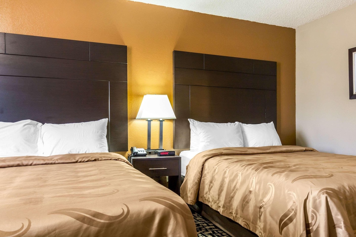 Quarto de hotel com duas camas, uma mesinha de cabeceira com abajur e um quadro pendurado na parede.
