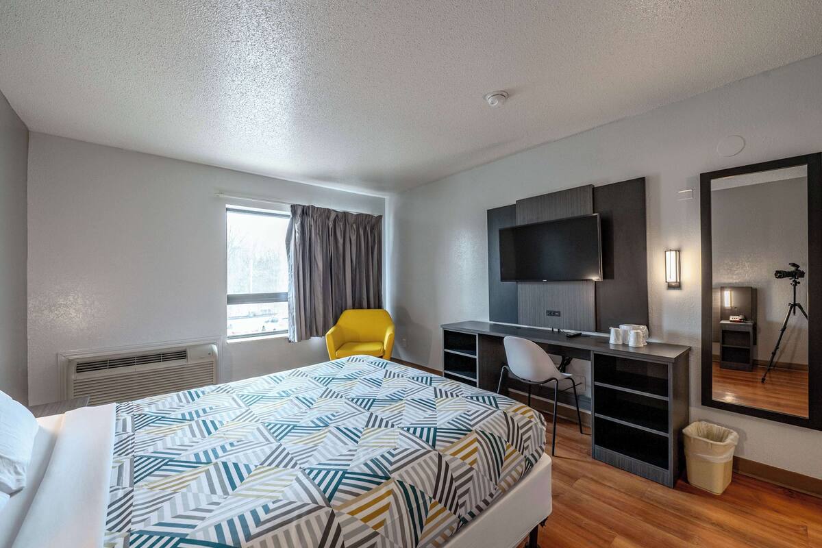 Une chambre d’hôtel comprenant un lit, un bureau avec une chaise, une télévision, un miroir et une fenêtre avec des rideaux.